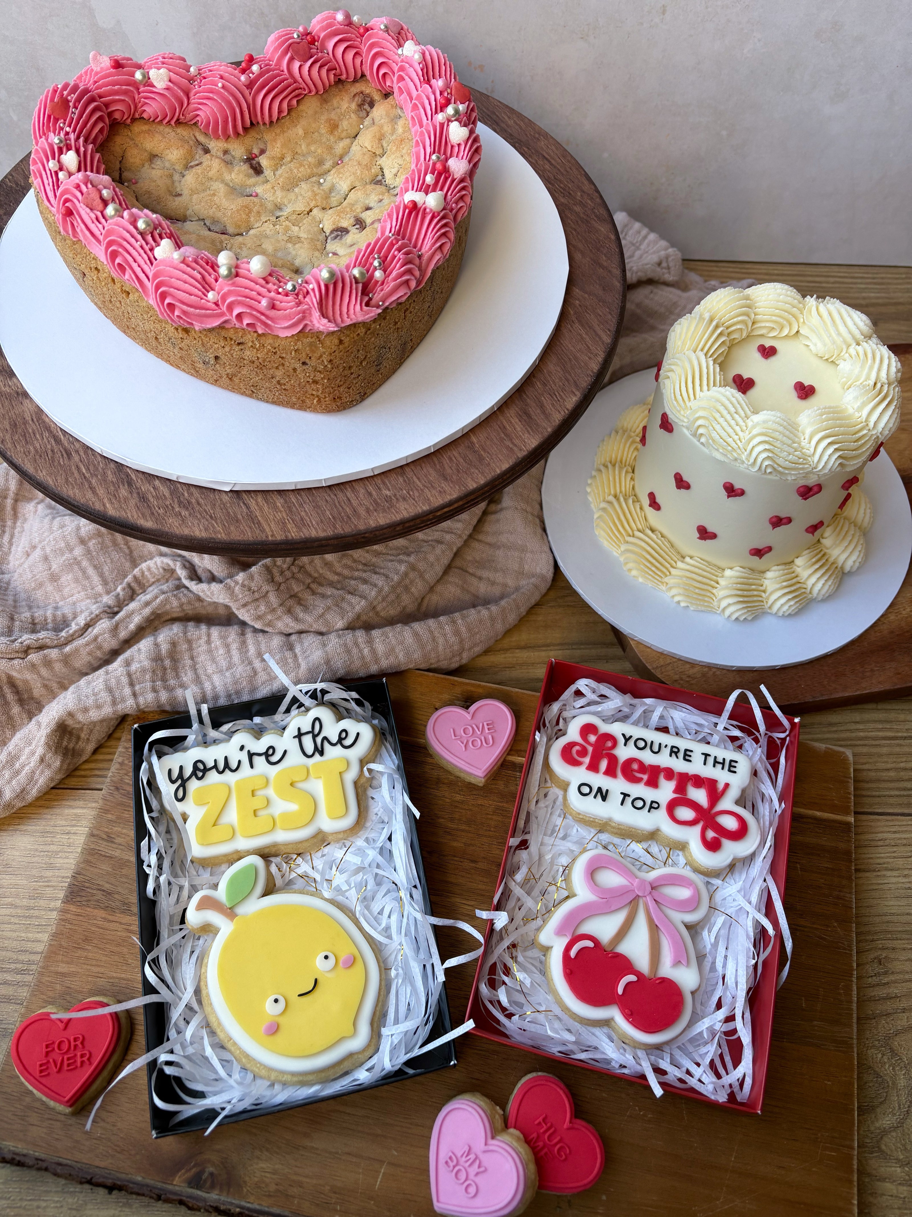 Heart-shaped cookie cake with pink frosting, a small decorated cake, and two cookie gift sets on a wooden surface.
